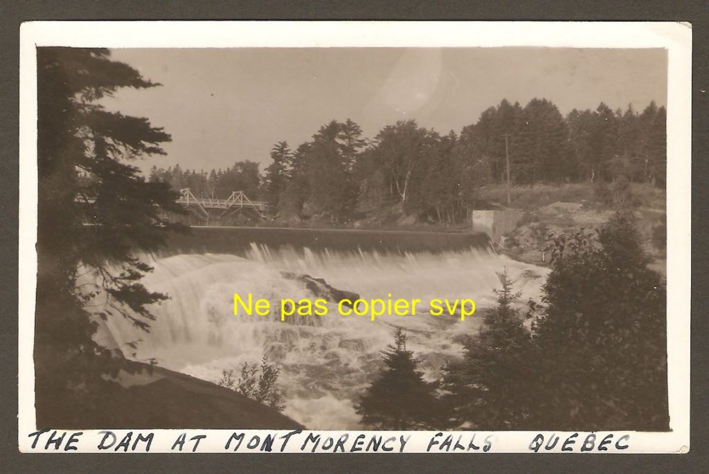 Tirage photographique de 1920 environ, montrant l'arête du barrage traversant la rivière Montmorency, juste avant le dénivelé de la chute. De plus, à l'arrière-plan, on remarque l'ancien pont qui enjambait alors la rivière. Au premier plan, à gauche, passe le conduit amenant l'eau de la station de pompage à la centrale hydro-électrique se trouvant en bas de la falaise, à l'ouest de la chute.